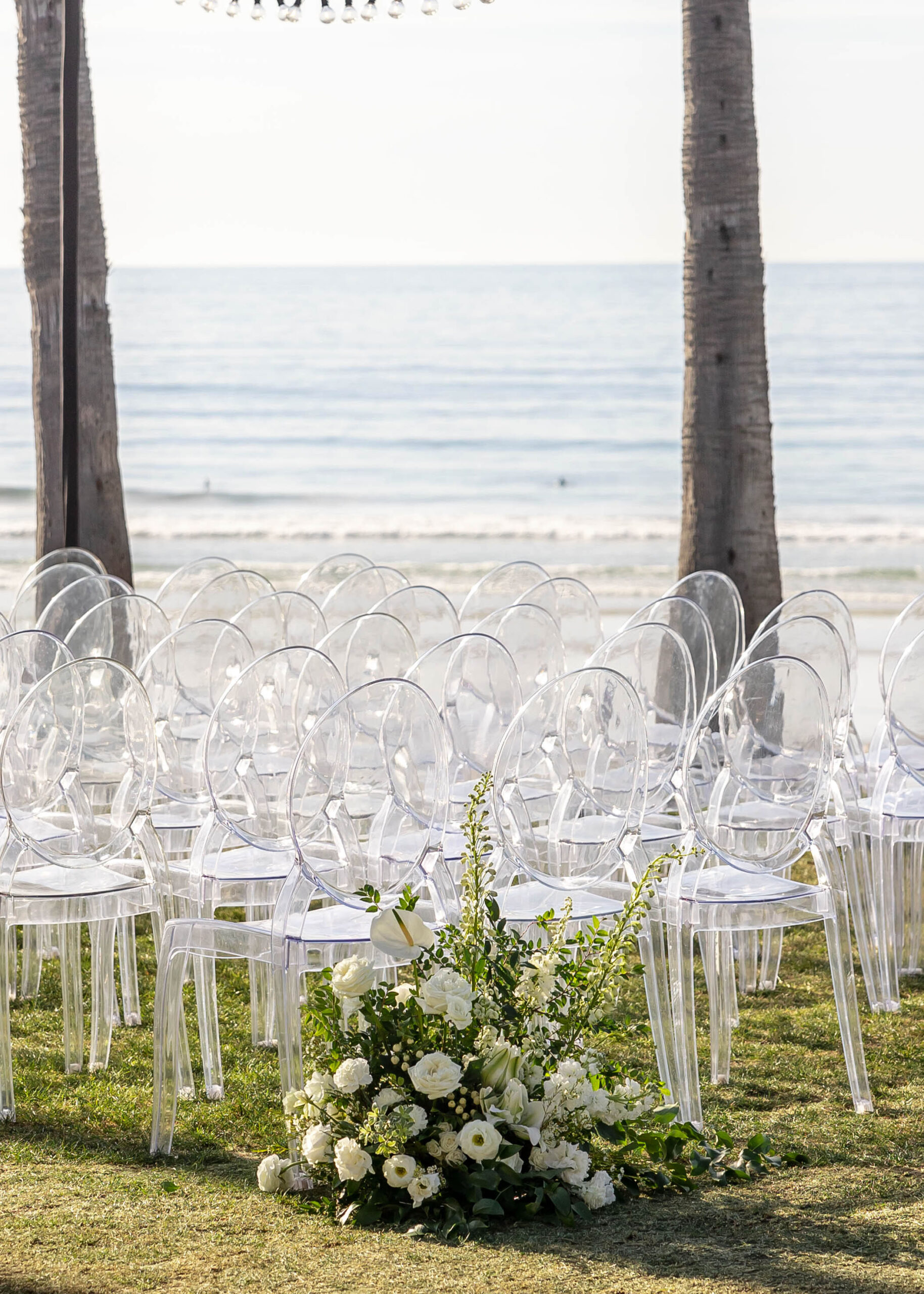 Beachside ceremony setup with clear chairs and white floral arch