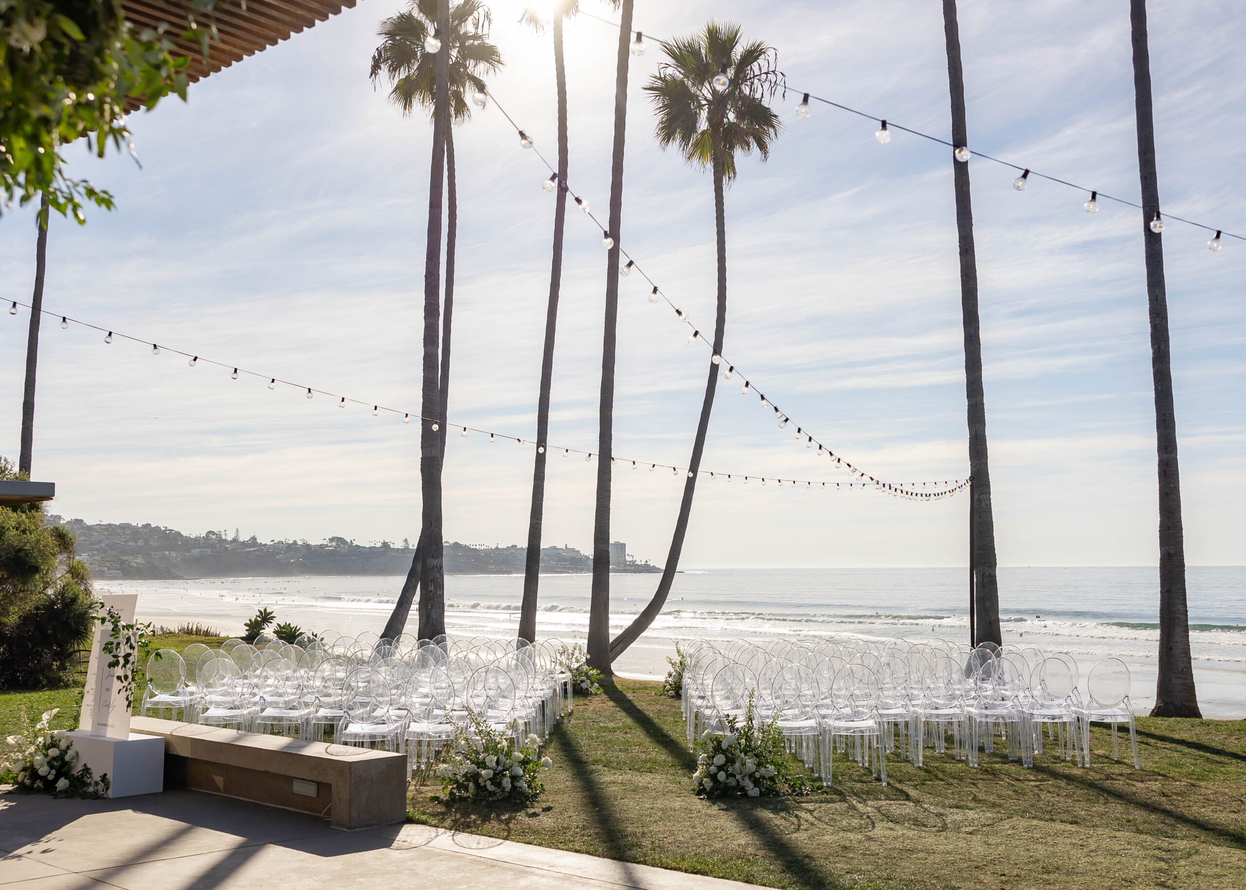 Beachside ceremony setup with clear chairs and white floral arch