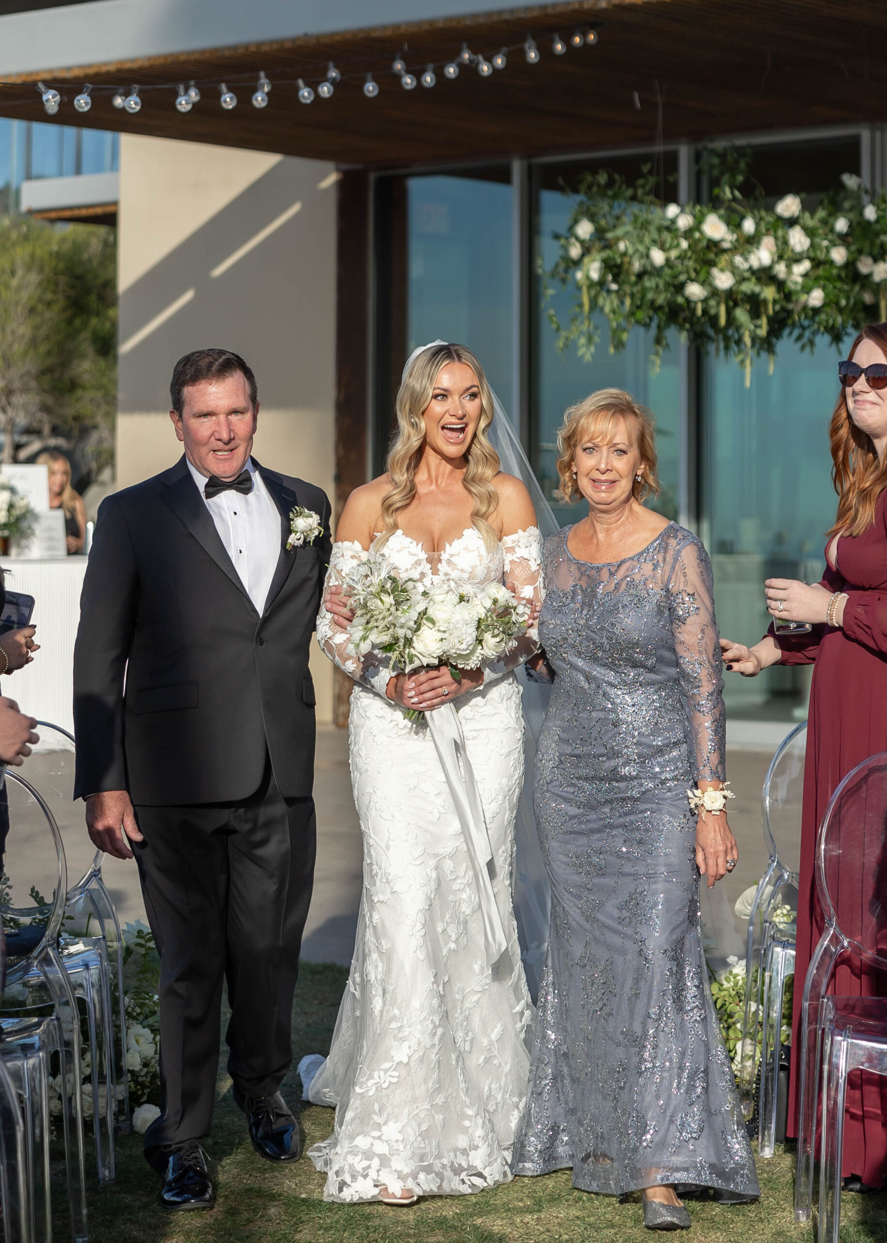 Beachside ceremony setup with clear chairs and white floral arch